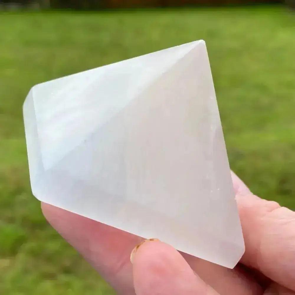 Selenite Crystal Destiny pyramid, luminous white and translucent, held in hand against green grass.
