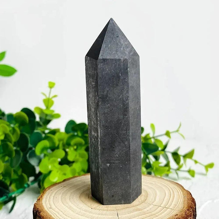 Dark gray shungite crystal tower from Crystal Destiny, on a wooden stand with green foliage.