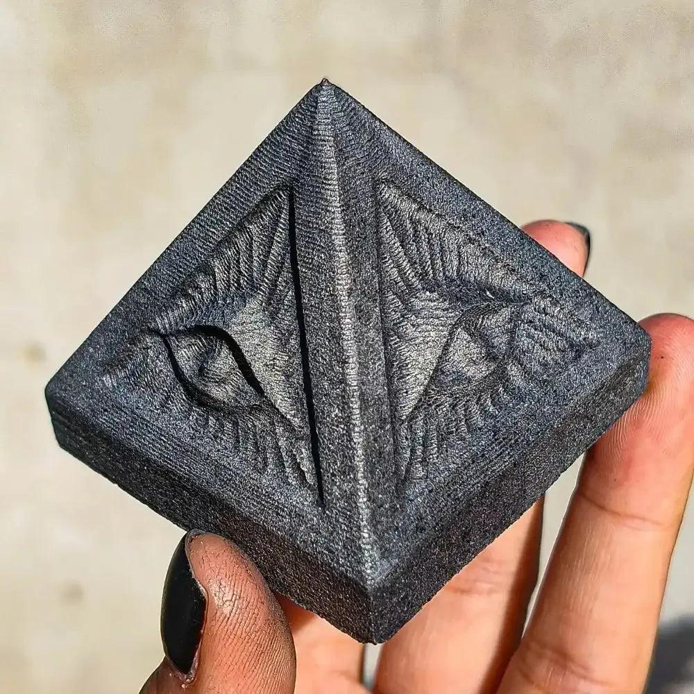 Hand-polished satin black Shungite pyramid with carved eye details for grounding, protection, and Root chakra healing.