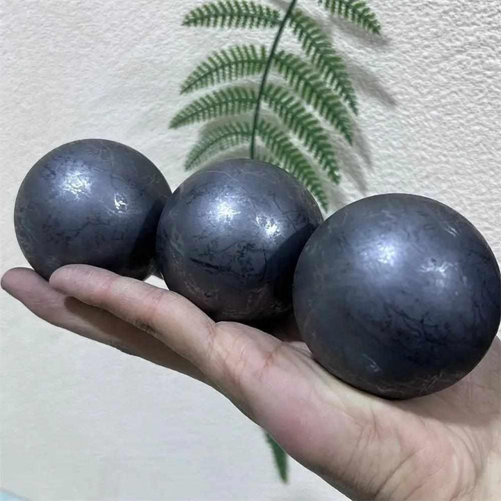 Three polished, deep black Shungite spheres with a metallic sheen, held in a hand for grounding and root chakra healing.