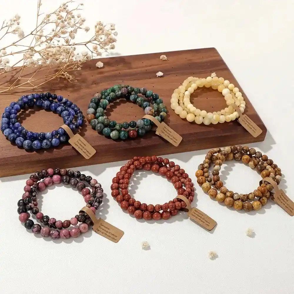 Six sets of natural stone beaded bracelets, including rhodonite, Indian agate, and other crystals, on a wooden board.