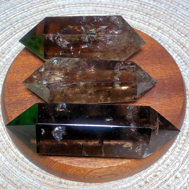 Three smoky quartz double point crystals with natural rainbow flash inclusions on a wooden display base.
