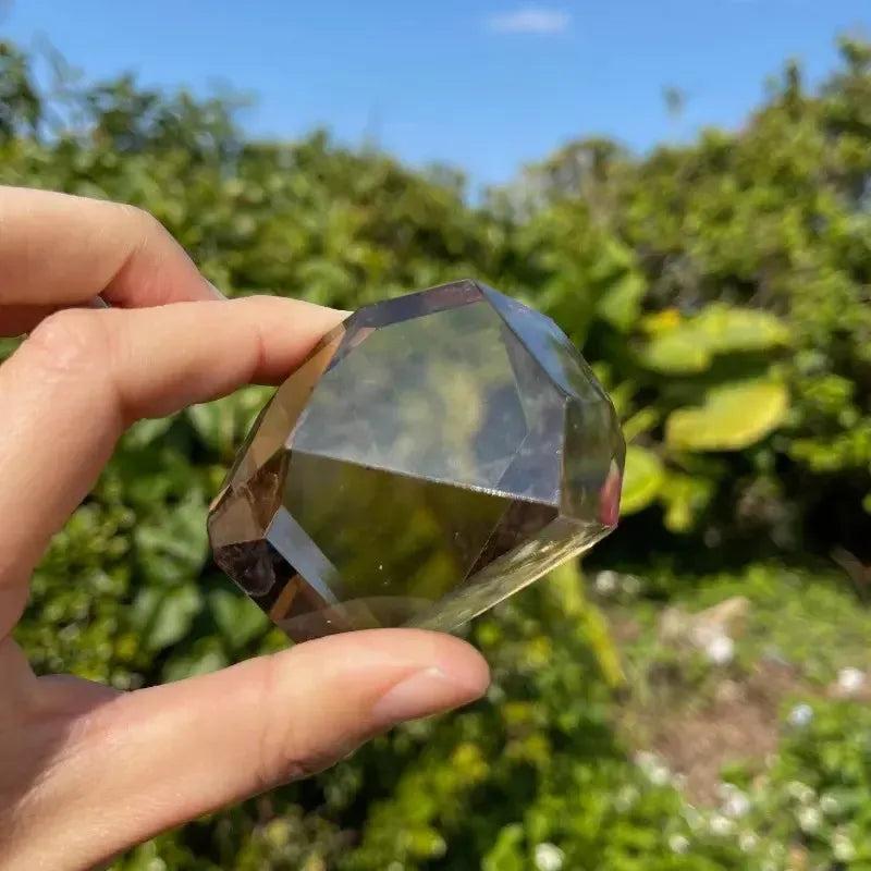 Hand holding a polished, faceted smoky quartz freeform crystal from Crystal Destiny, against a green outdoor background.