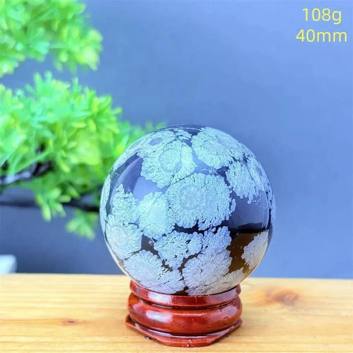 Snowflake Obsidian Sphere: Polished black volcanic crystal with striking white snowflake patterns on a wooden stand.