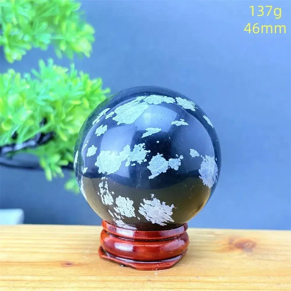 Snowflake Obsidian Sphere: Polished black volcanic crystal with striking white snowflake patterns on a wooden stand.