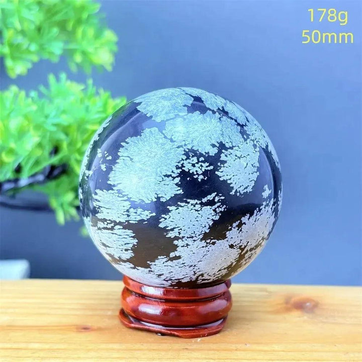 Snowflake Obsidian Sphere: Deep black volcanic crystal with striking white snowflake patterns on a wooden stand.