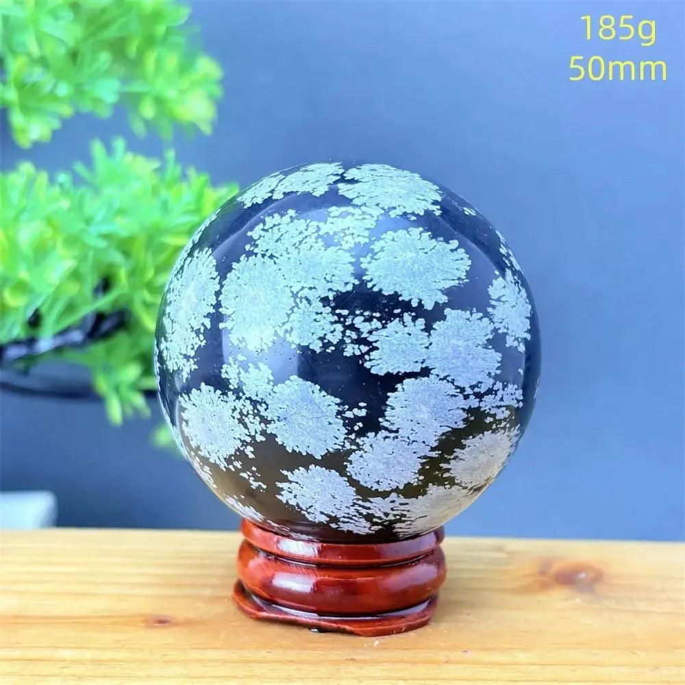Snowflake Obsidian Sphere: Deep black volcanic crystal with striking white snowflake patterns on a wooden stand.