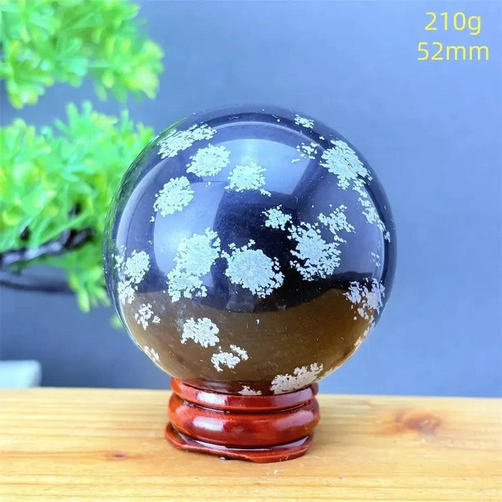 Snowflake Obsidian Sphere: Polished black volcanic crystal with striking white snowflake patterns on a wooden stand.