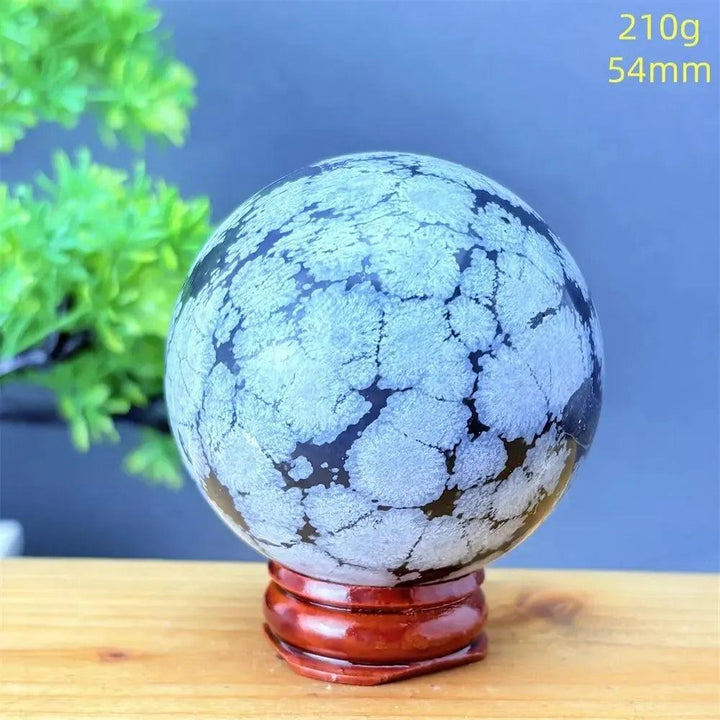 Snowflake Obsidian Sphere: Deep black volcanic crystal with striking white snowflake patterns on a wooden stand.