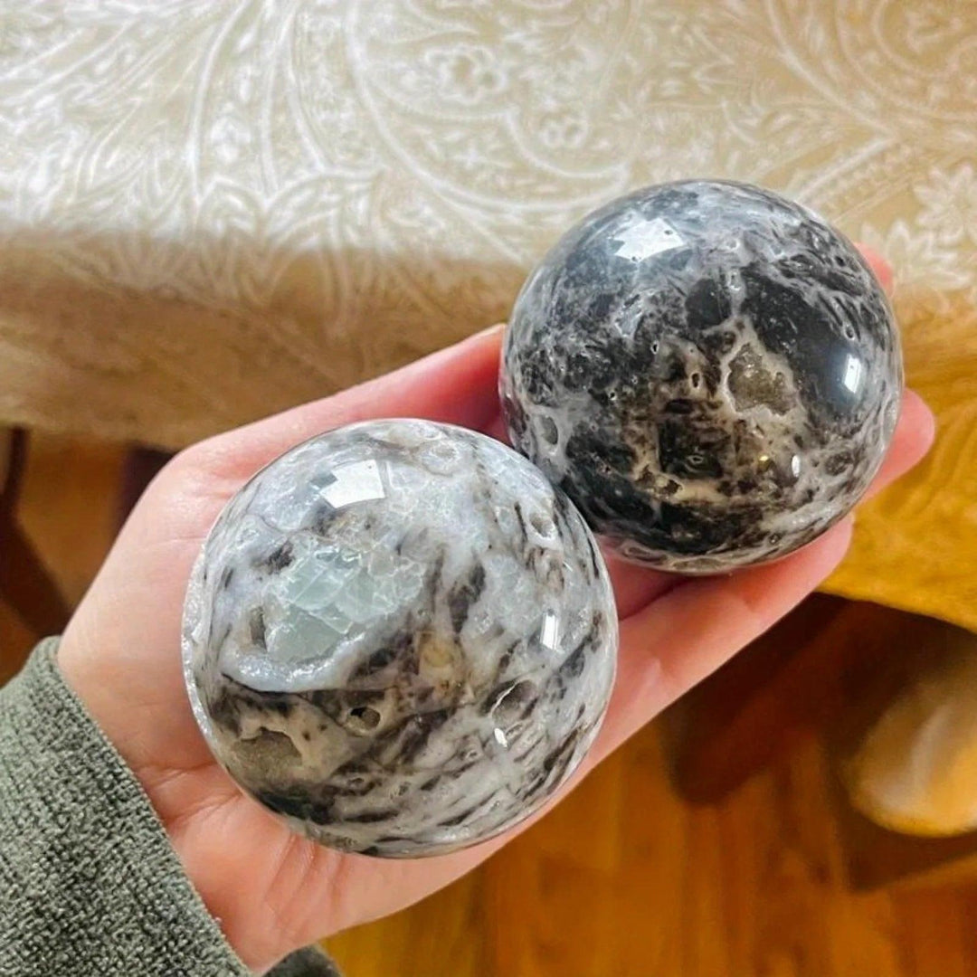 Two polished gray and black Sphalerite crystal spheres with intricate patterns, held in a hand.