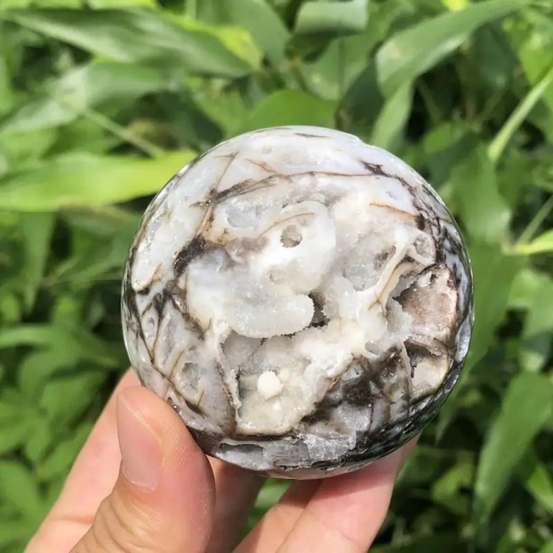 Unique gray and black Sphalerite crystal sphere with intricate patterns and white druzy, held against green foliage.