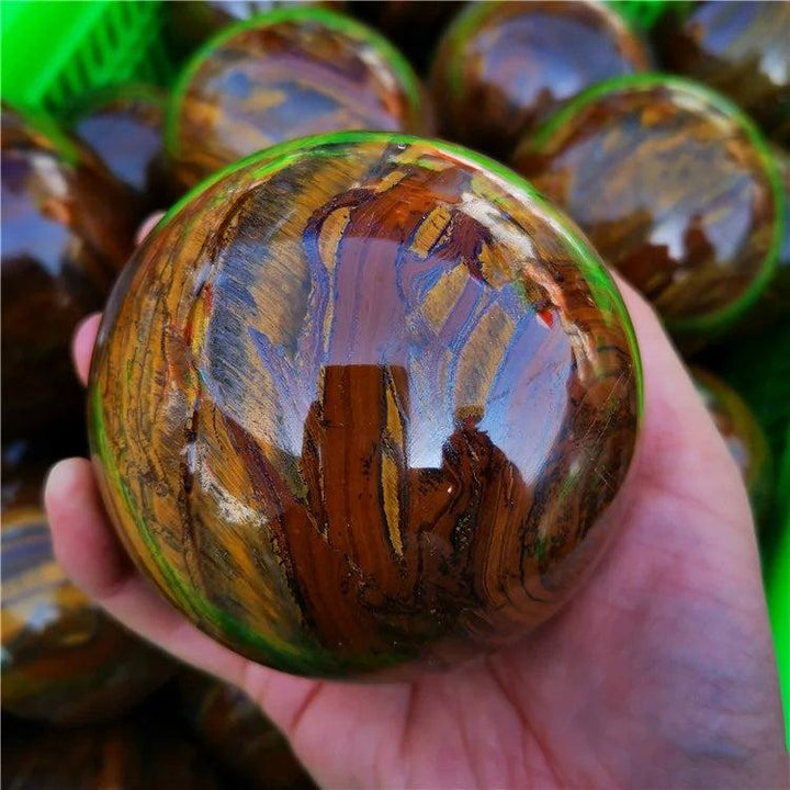 Polished Tiger Iron crystal sphere with golden shimmer, deep red, and metallic silver bands, held in a hand.