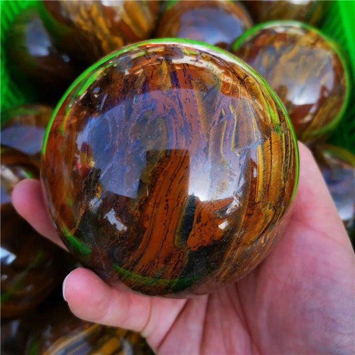 Polished Tiger Iron crystal sphere with golden shimmer, deep red, and metallic silver bands, held in a hand.