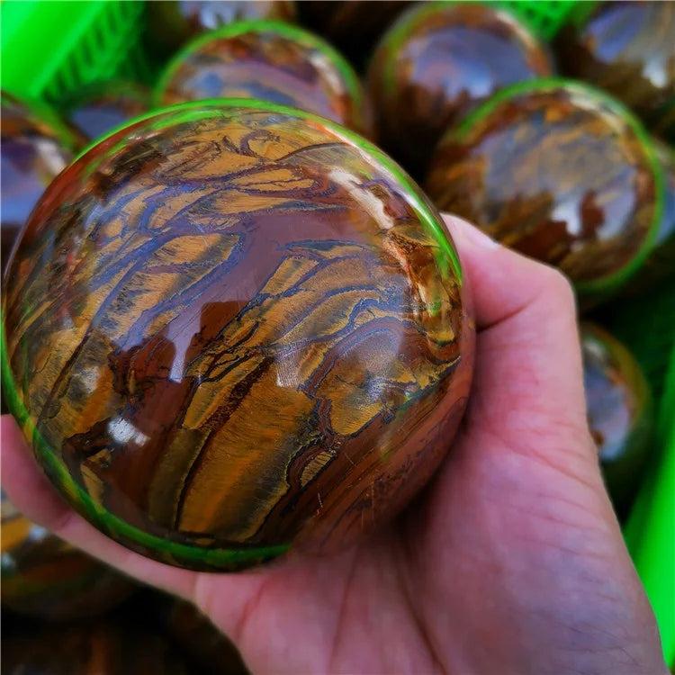 Hand holding a polished Tiger Iron crystal sphere with golden shimmer, deep red, and metallic silver bands.