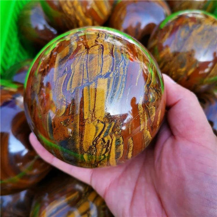 Hand holding a polished Tiger Iron crystal sphere with golden shimmer, deep red, and metallic silver bands.