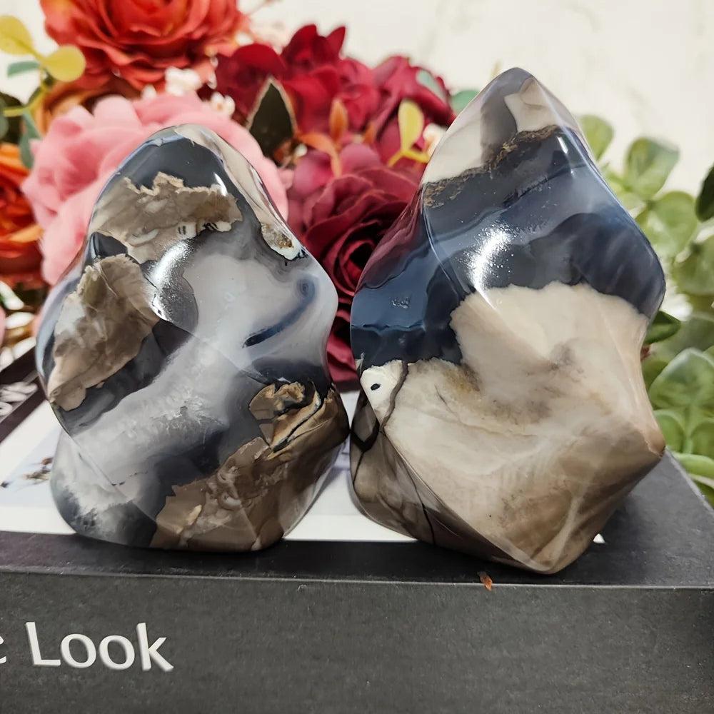 Two polished volcanic agate flame crystals with swirling charcoal, cream, and brown patterns, set against flowers.