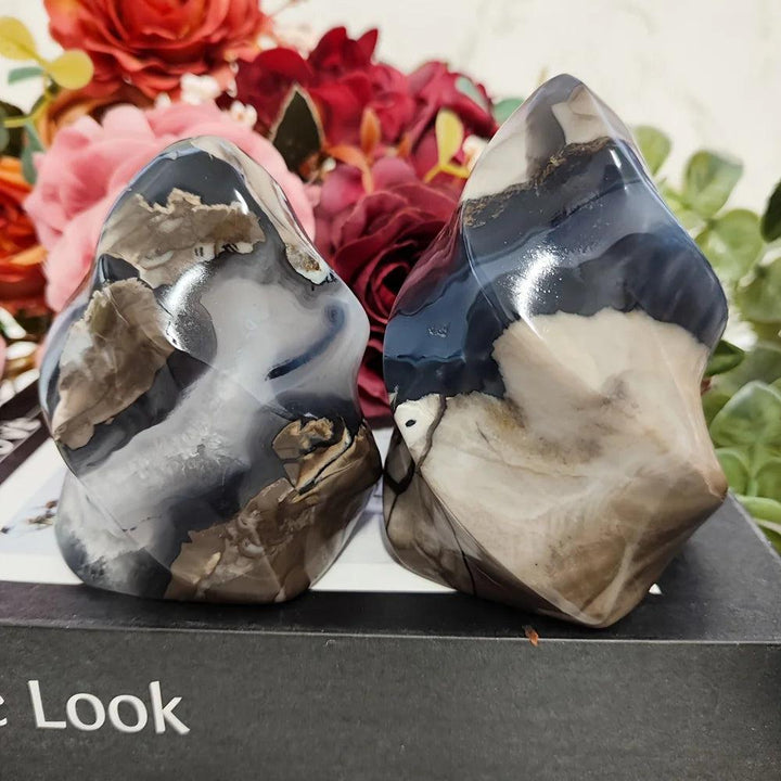 Two polished volcanic agate flame crystals with swirling charcoal, cream, and brown patterns, set against flowers.