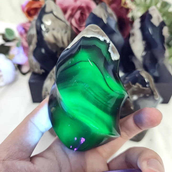 Hand holding a glowing green UV reactive volcanic agate flame crystal, with other crystals in background.