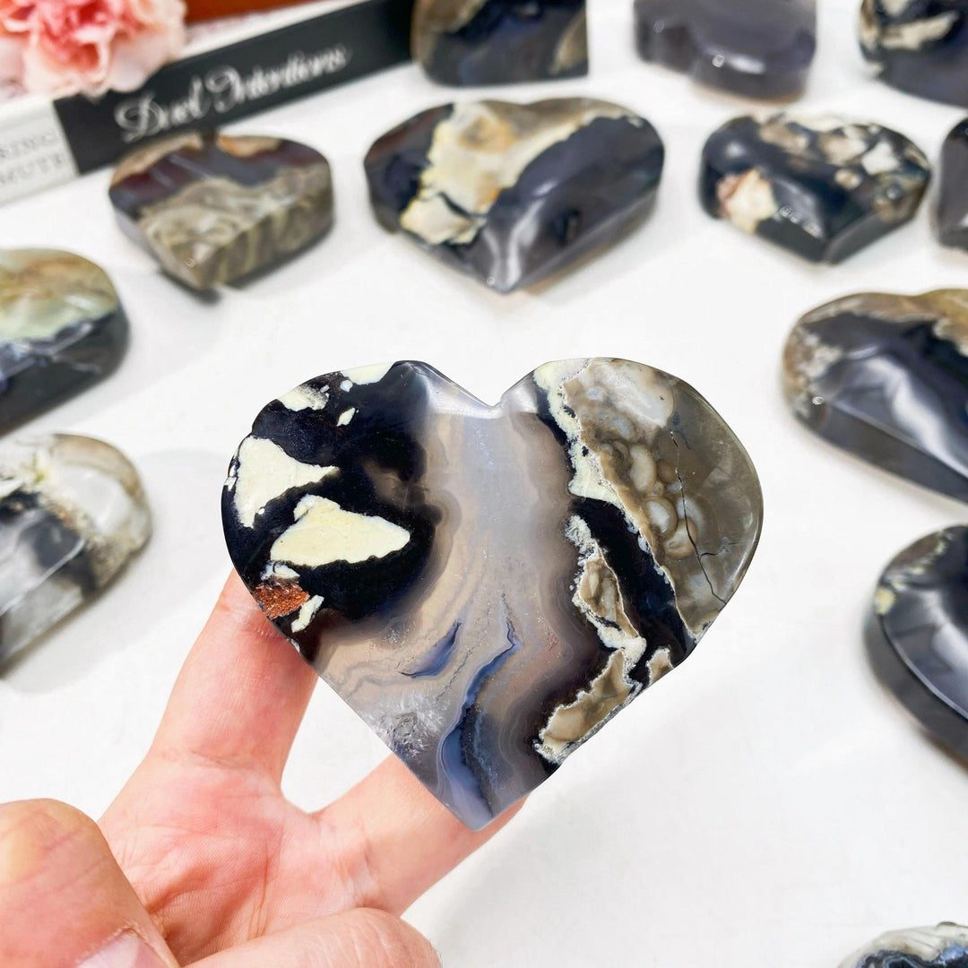 Hand holding a polished volcanic agate heartstone with swirling black, white, and gray patterns.