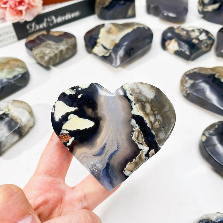 Hand holding a polished volcanic agate heartstone with swirling black, white, and gray patterns.