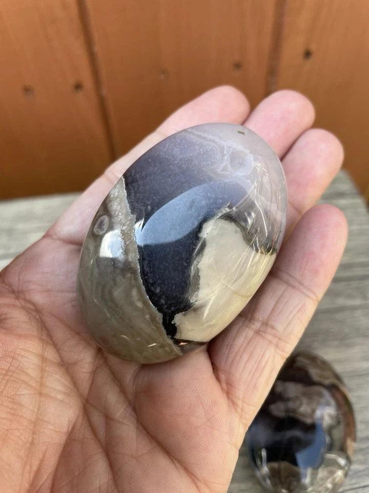 Smooth, polished natural volcanic agate palm stone with swirling grey, black, and white patterns, held in a hand.