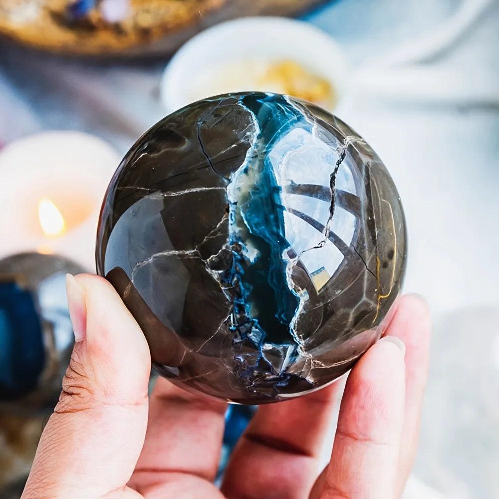 Hand holding a polished volcanic agate sphere with dark charcoal, cream, and blue-green veins, reflecting light.