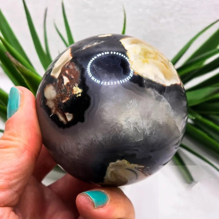 Polished volcanic agate sphere, 50-80mm, with charcoal, cream, and earthy tones, held by a hand.