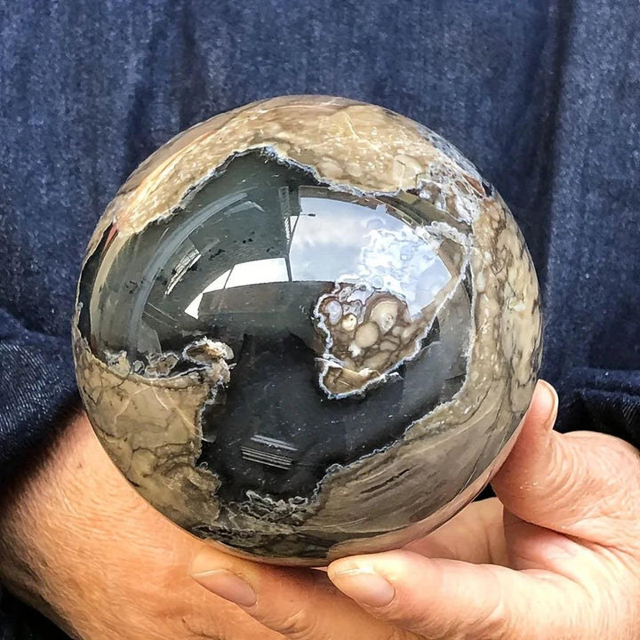 Polished volcanic agate sphere, 50-80mm, with swirling charcoal, cream, and earthy tones, held in hands.