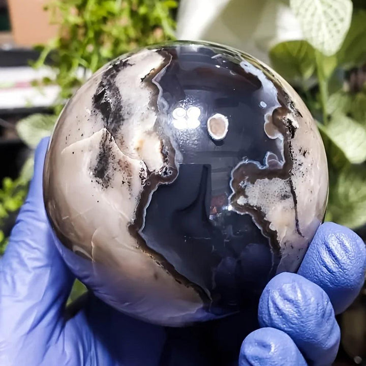 Polished volcanic agate sphere, 50-80mm, with swirling charcoal, cream, and earthy tones, held in a gloved hand.