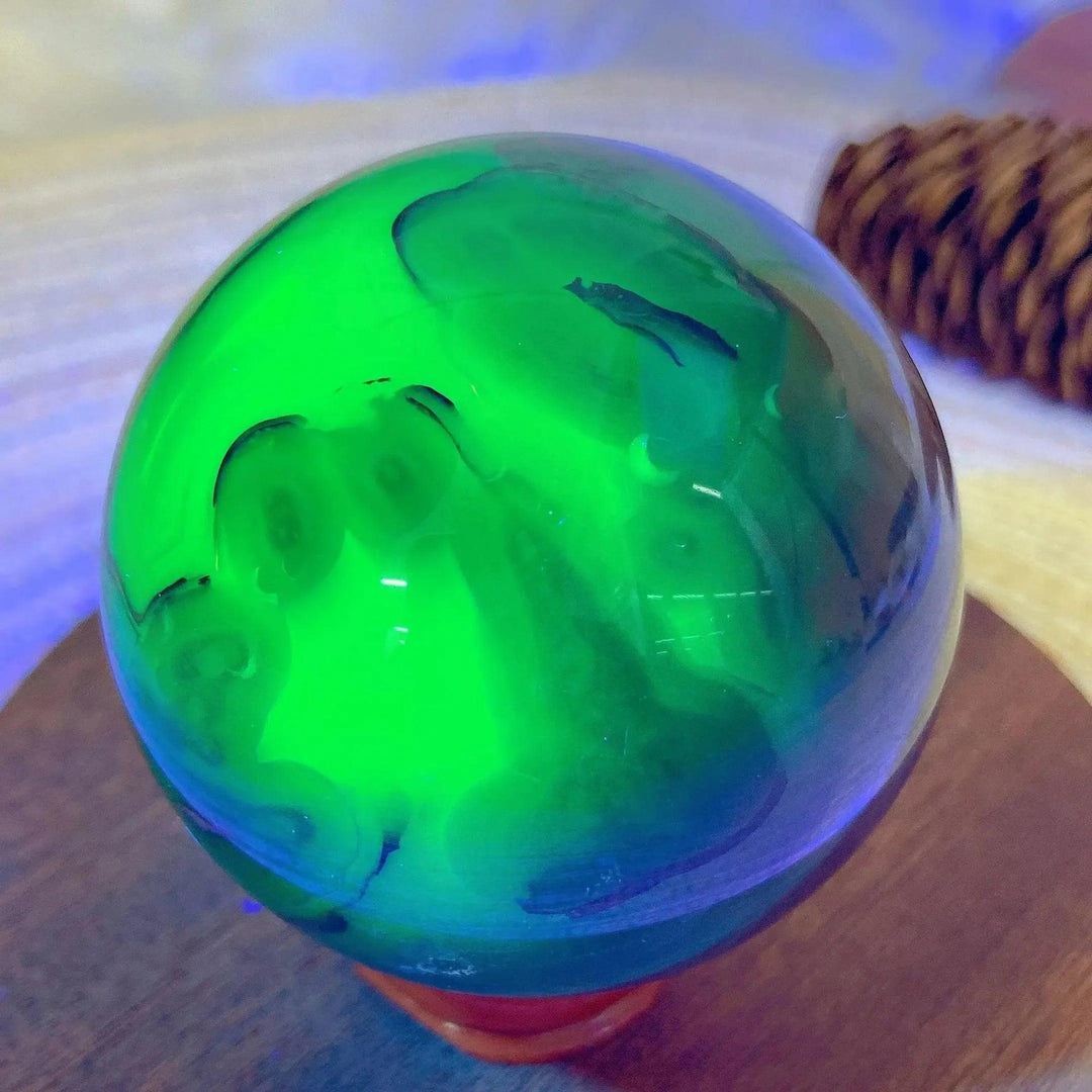 Volcanic Agate Sphere, a UV reactive crystal ball, glowing vibrant green with dark blue swirls, on a wooden base.