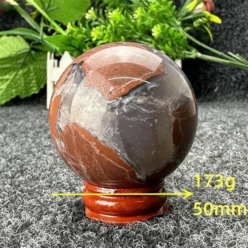 Polished Volcano Agate sphere from Crystal Destiny, displaying dramatic brown, red, and gray patterns on a wooden stand.