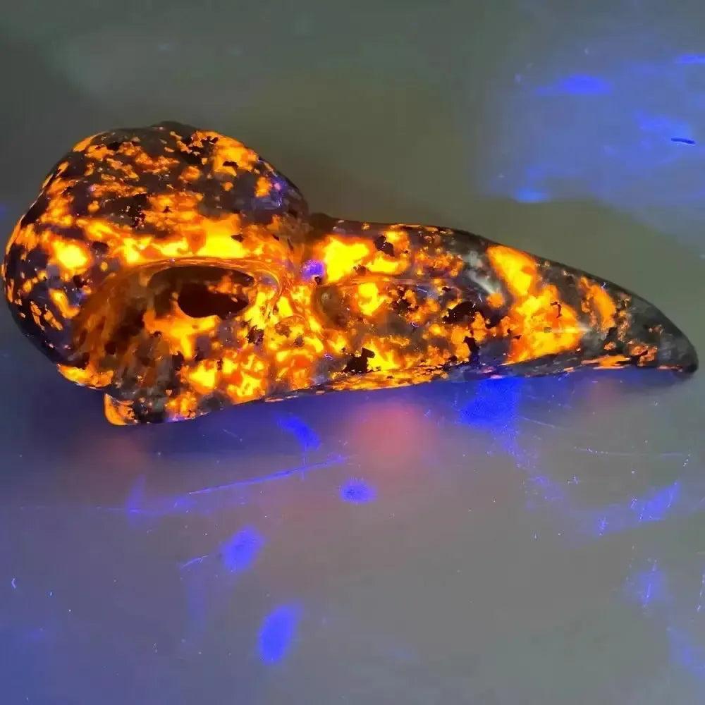 Yooperlite Crow Skull Crystal Carving, UV reactive, glowing vivid orange and yellow, a talisman of mystery and transformation.