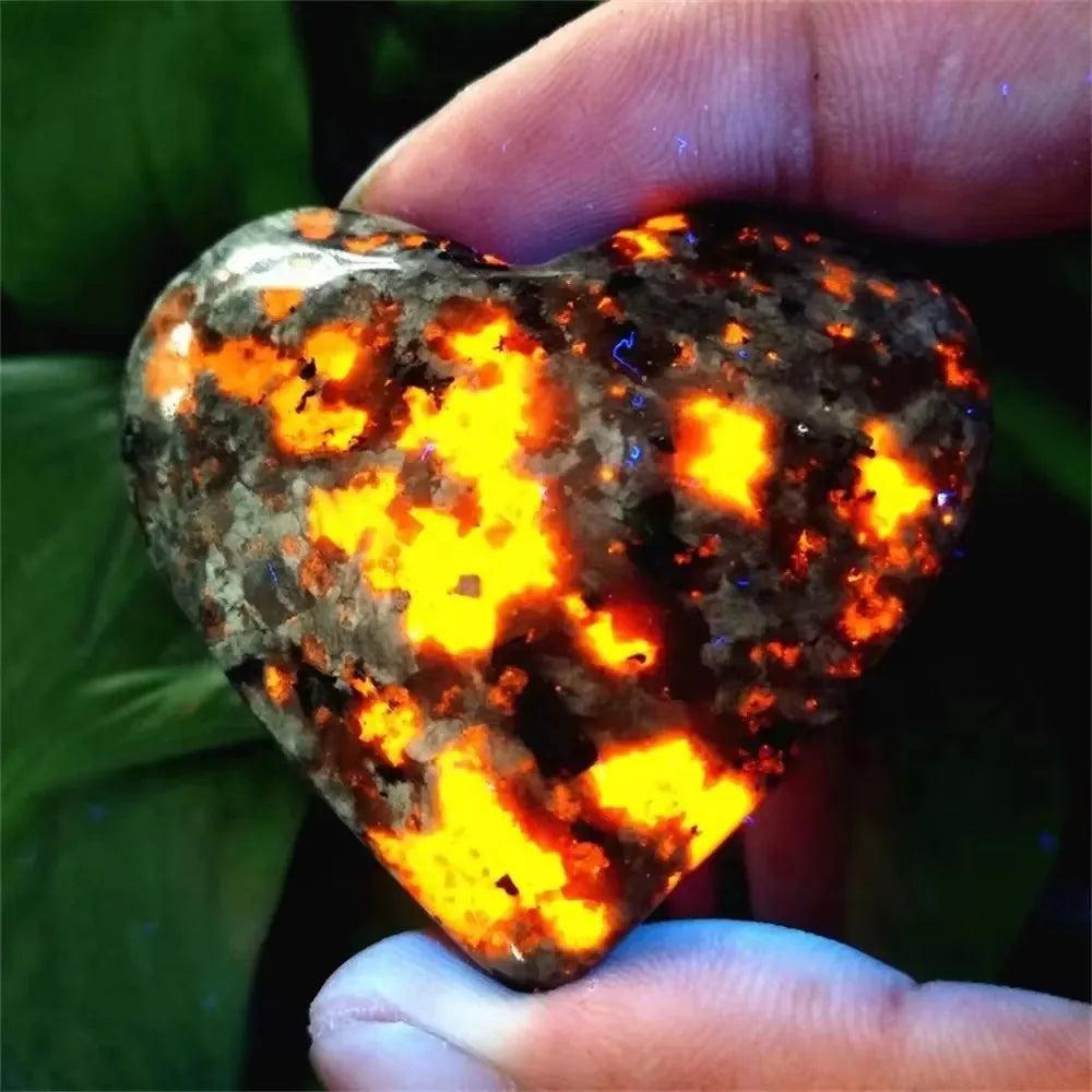 Yooperlite Crystal Heart Stone glowing vivid orange under UV light, held by fingers. Supports Third Eye, Root, Sacral chakras.