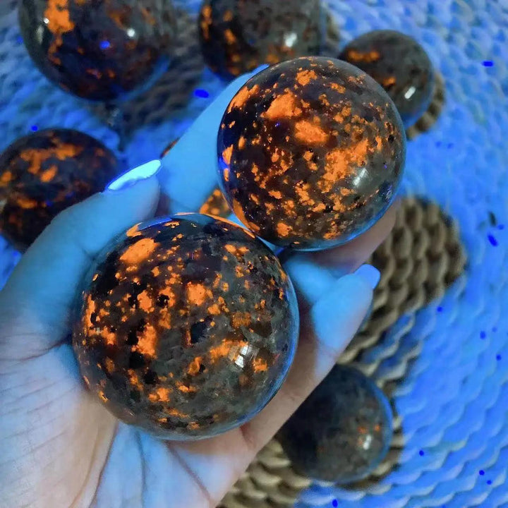 Hand holding two Yooperlite Crystal Sphere Flame UV Reactive Glowing Balls, vivid orange and gold patterns on dark stone.
