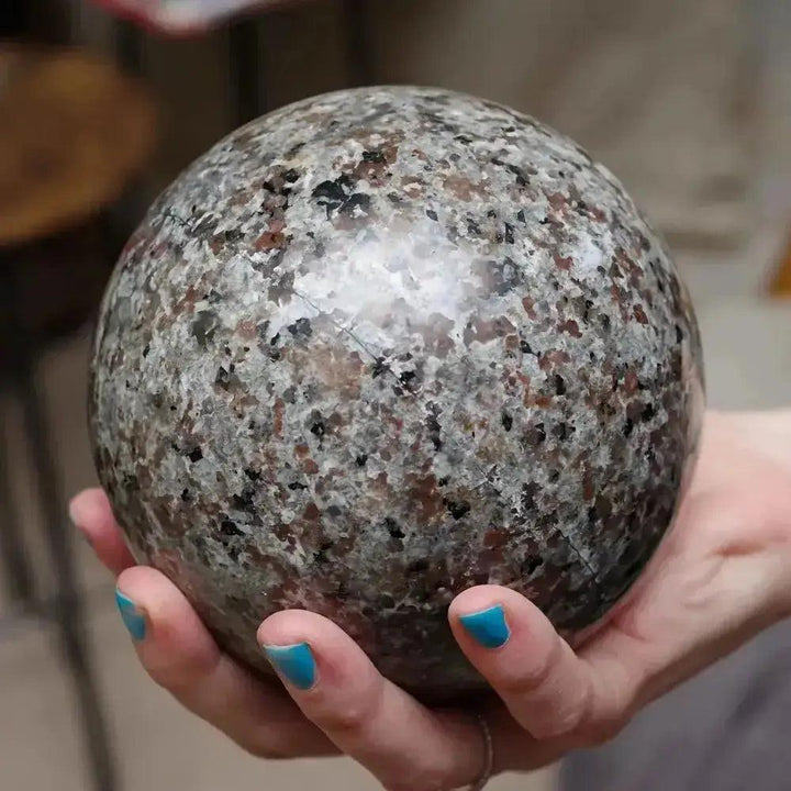 Yooperlite UV Glowing Crystal Sphere, a marbled gray sphere with black and reddish flecks, held in a hand.