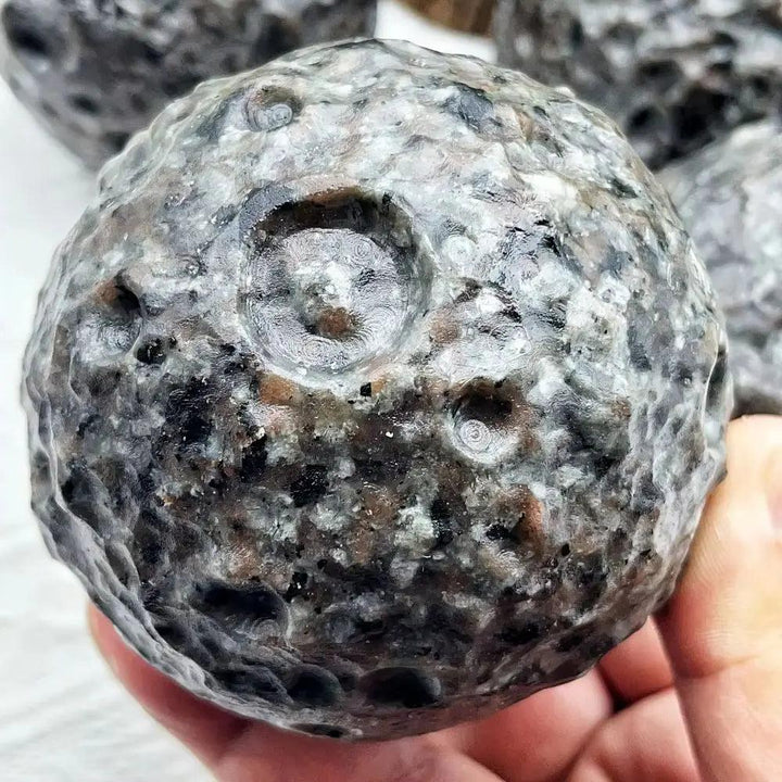 Hand holding a grey Yooperlite Crystal Sphere, a UV reactive glowing moon ball with unique crater-like patterns.
